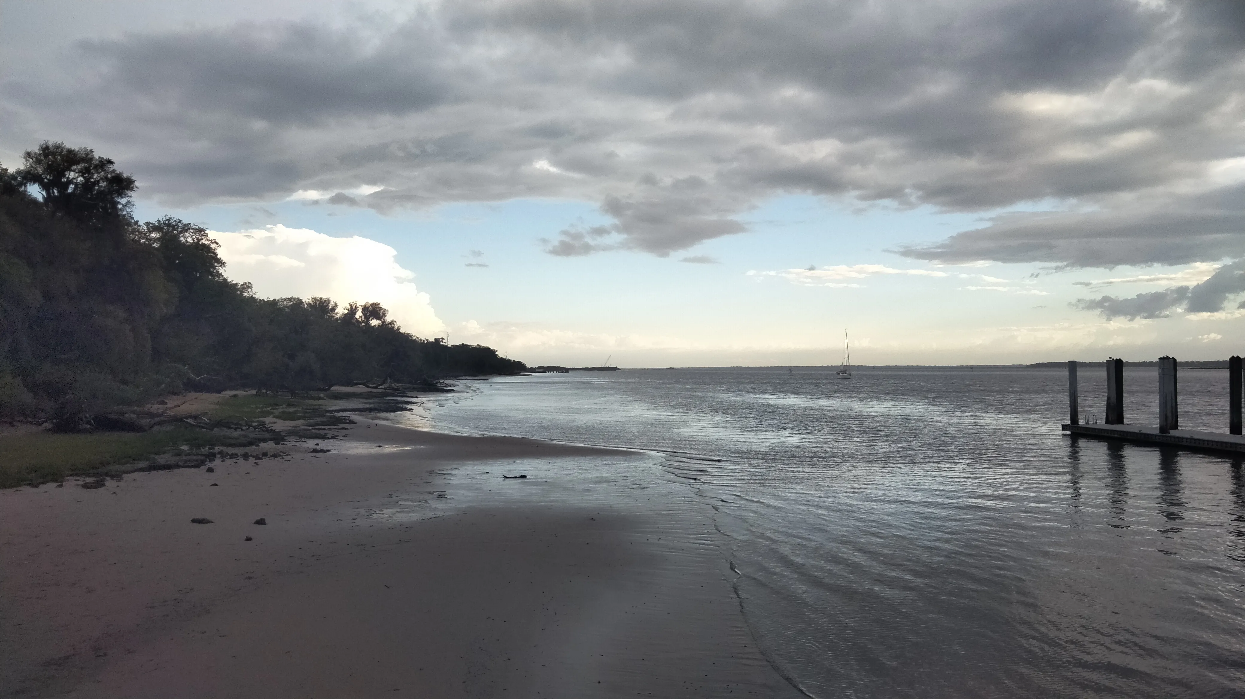 A river with a sailboat off center to the right. A large storm cloud is visible in the distance at sunset.