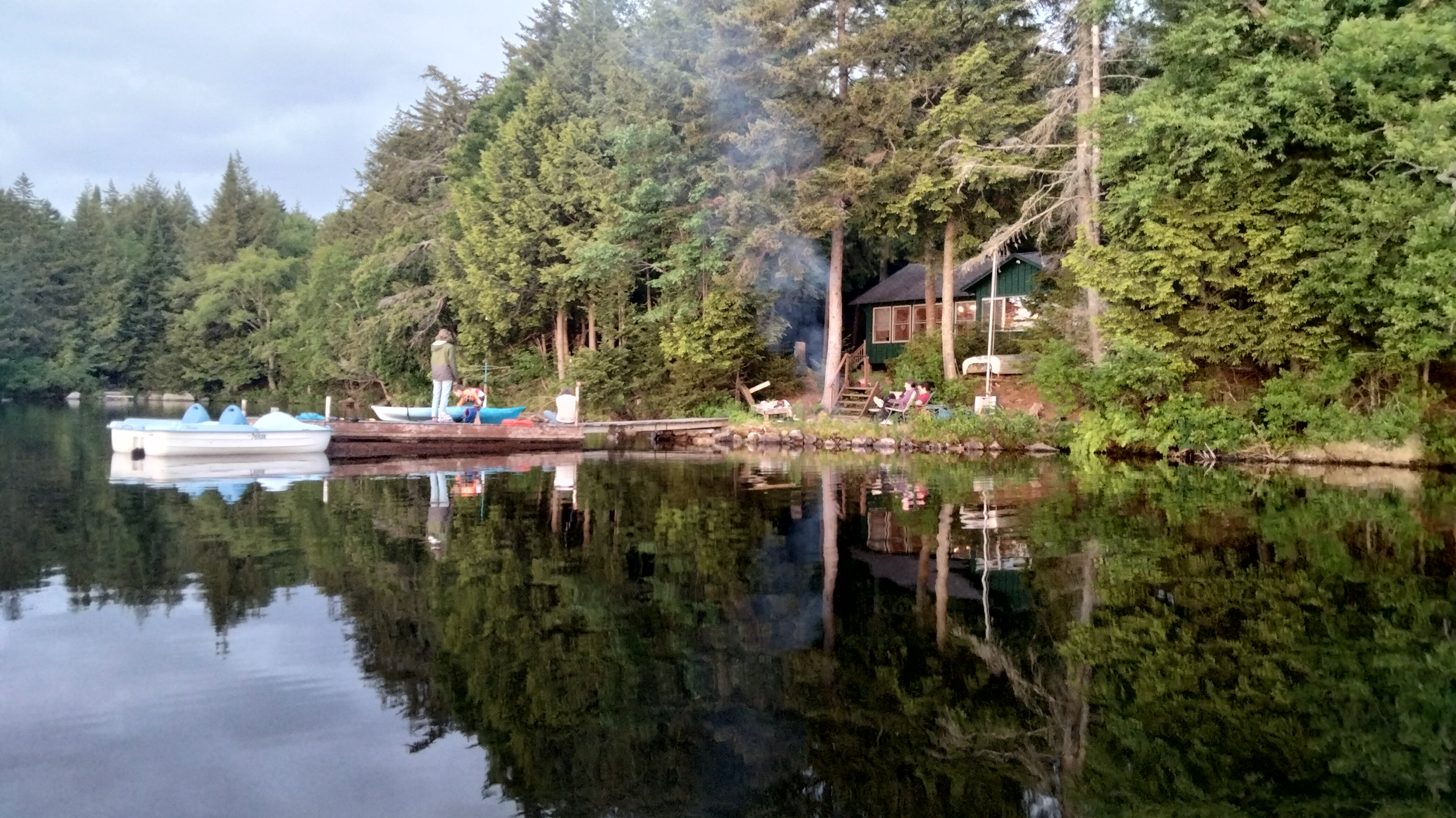 A dock, cabin, and fire taken from a canoe. A woman in a green raincoat is standing on the dock talking to kids near a kayak. Two men are sitting near a small camp fire on shore.
