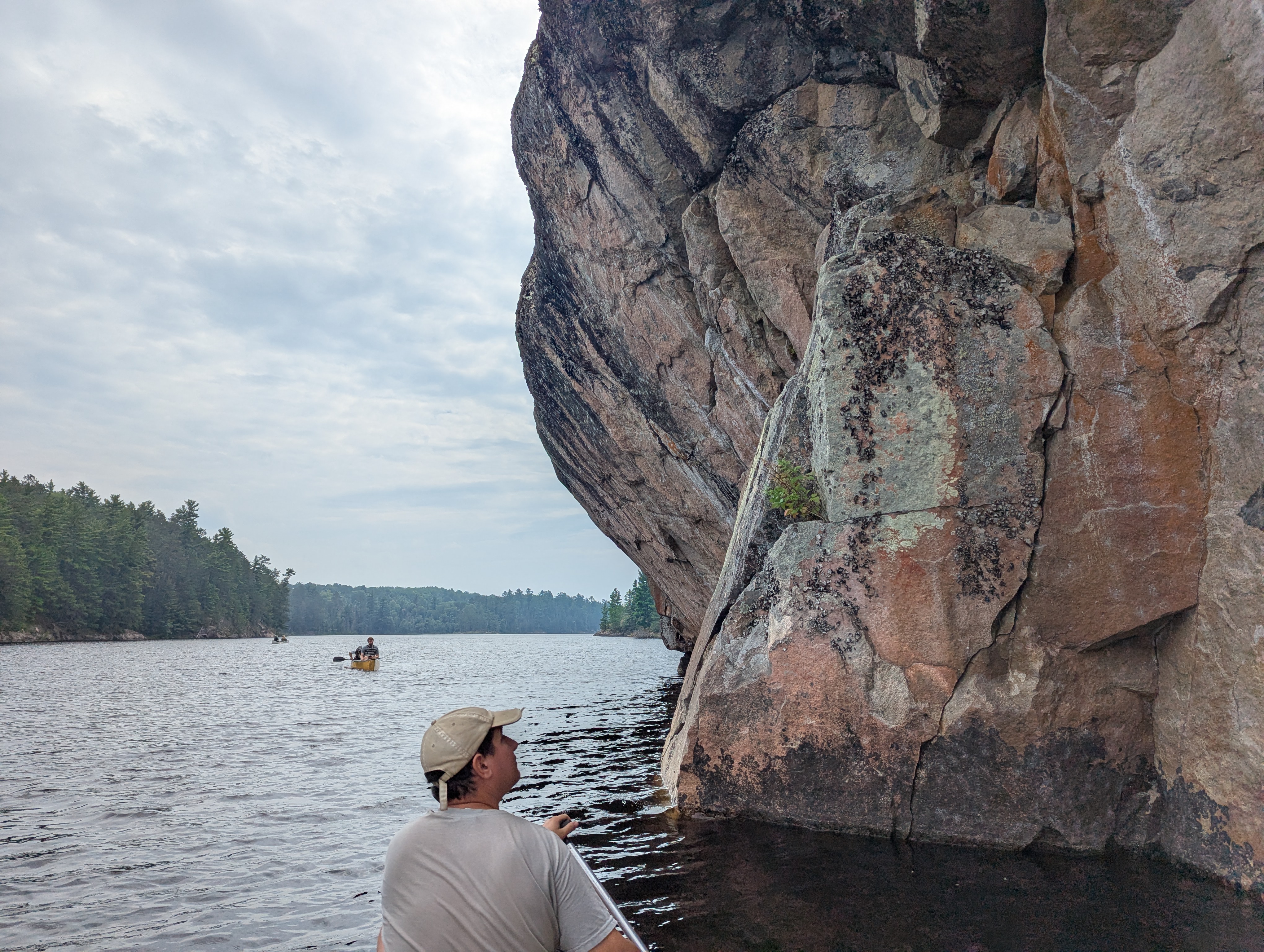 A man sits in a canoe looking at a sheer cliff on his right. A rock juts out over the water at a sharp angle.