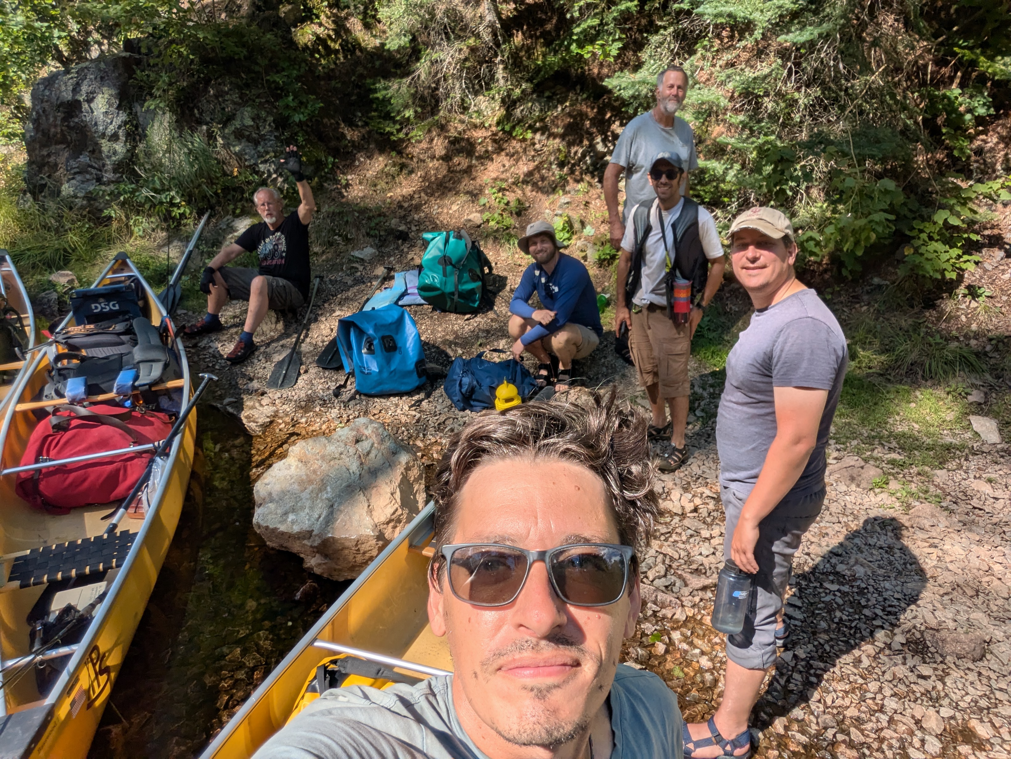 A group of men in a selfie photo. Canoes and camping packs are also in the picture. We are resting on the shore after a portage.
