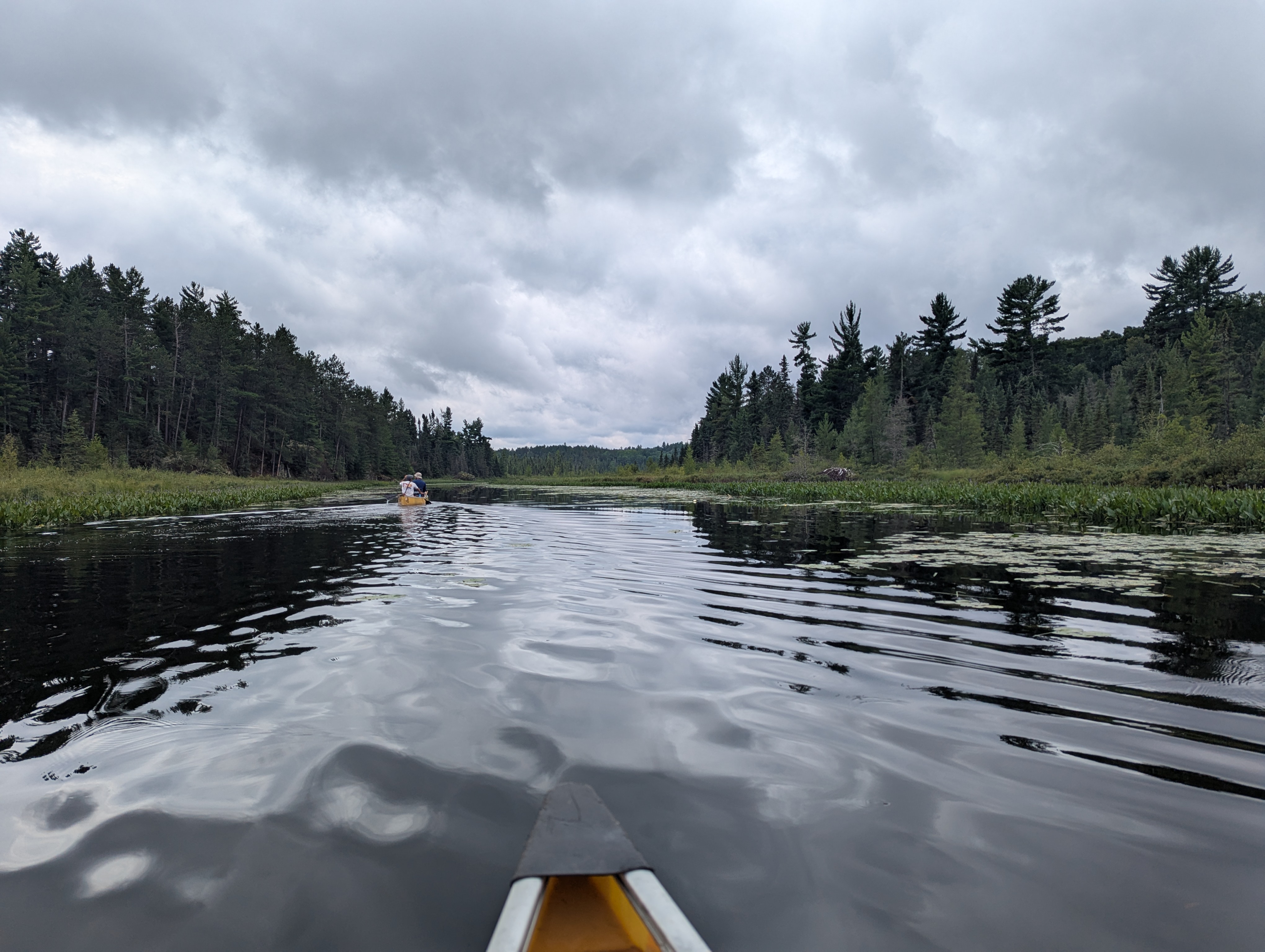 A canoe prow is visible on ripply water. Trees border the distance. Another canoe is visible ahead.