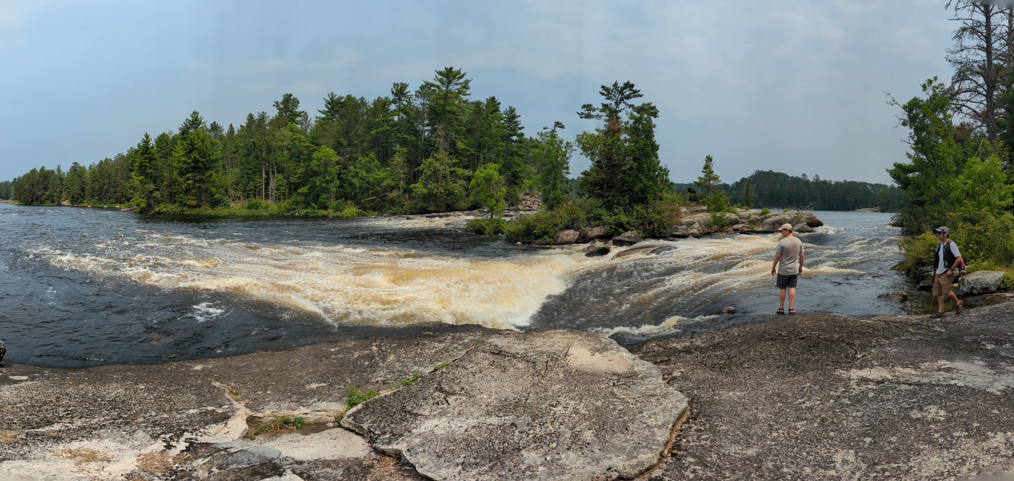 A panoramic photograph of a waterfall flowing right to left. A large rock face has people looking at the water in the foreground.