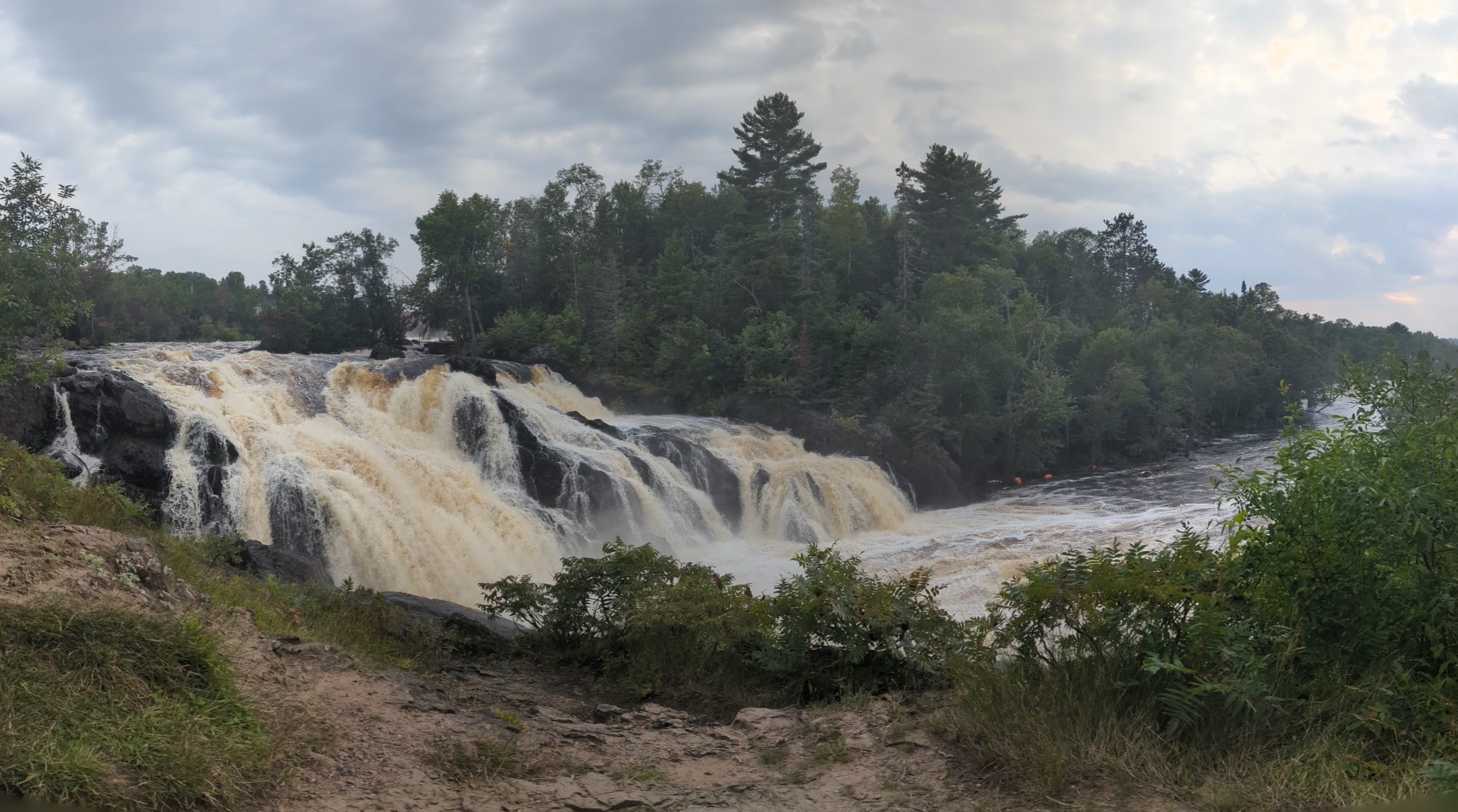 A panoramic photograph of a waterfall. A large waterfall on the left flows violently to a lake in the distance on the right.