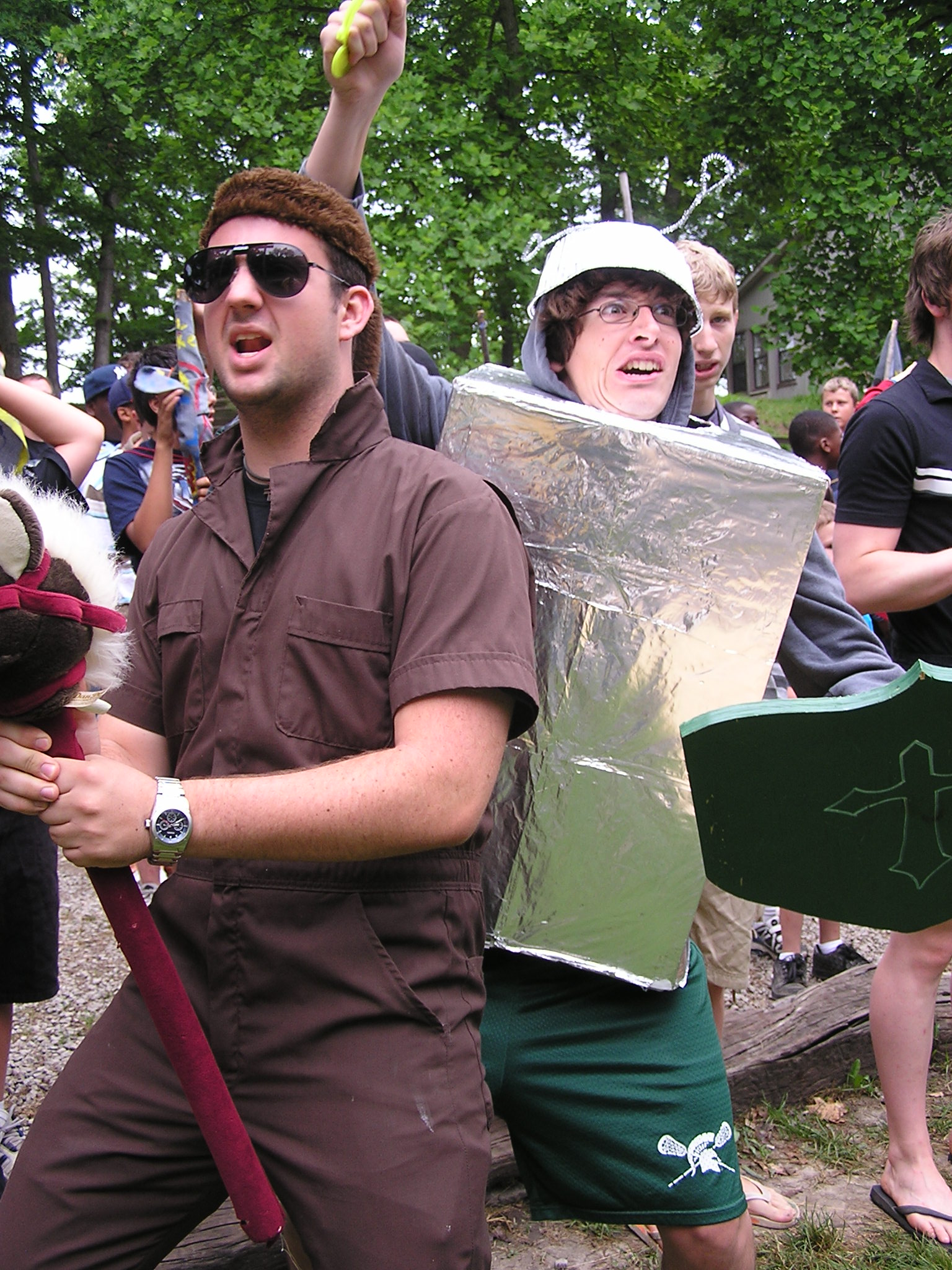 Two men. One dressed in a brown coverall wearing aviator sunglasses and a coonskin cap holding a toy horse - the kind with a horse head on a stick. He is yelling something. Another man wearing a robot costume that is supposed to look like armor. He is making a savage face and weilding a toy sword and shield. Togteher, they are Don Quixote and his horse, Rocinante.