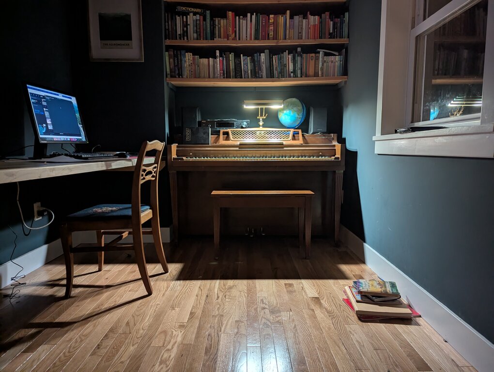 A piano under a bookshelf lit by a single light. A computer monitor is on the desk to the left, a chair slightly pulled out. It is a very dark picture, the only light coming from the lamp on the piano.