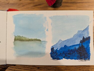 Two small landscapes. The left is a peninsula of trees in the distance with water in the foreground. The trees are lightly reflected in the water. The right is a mountainscape in all blues. A light blue sky is above pale blue mountains which are covered by a much darker blue range in the foreground. Black pen evergreen trees are also in the foreground.