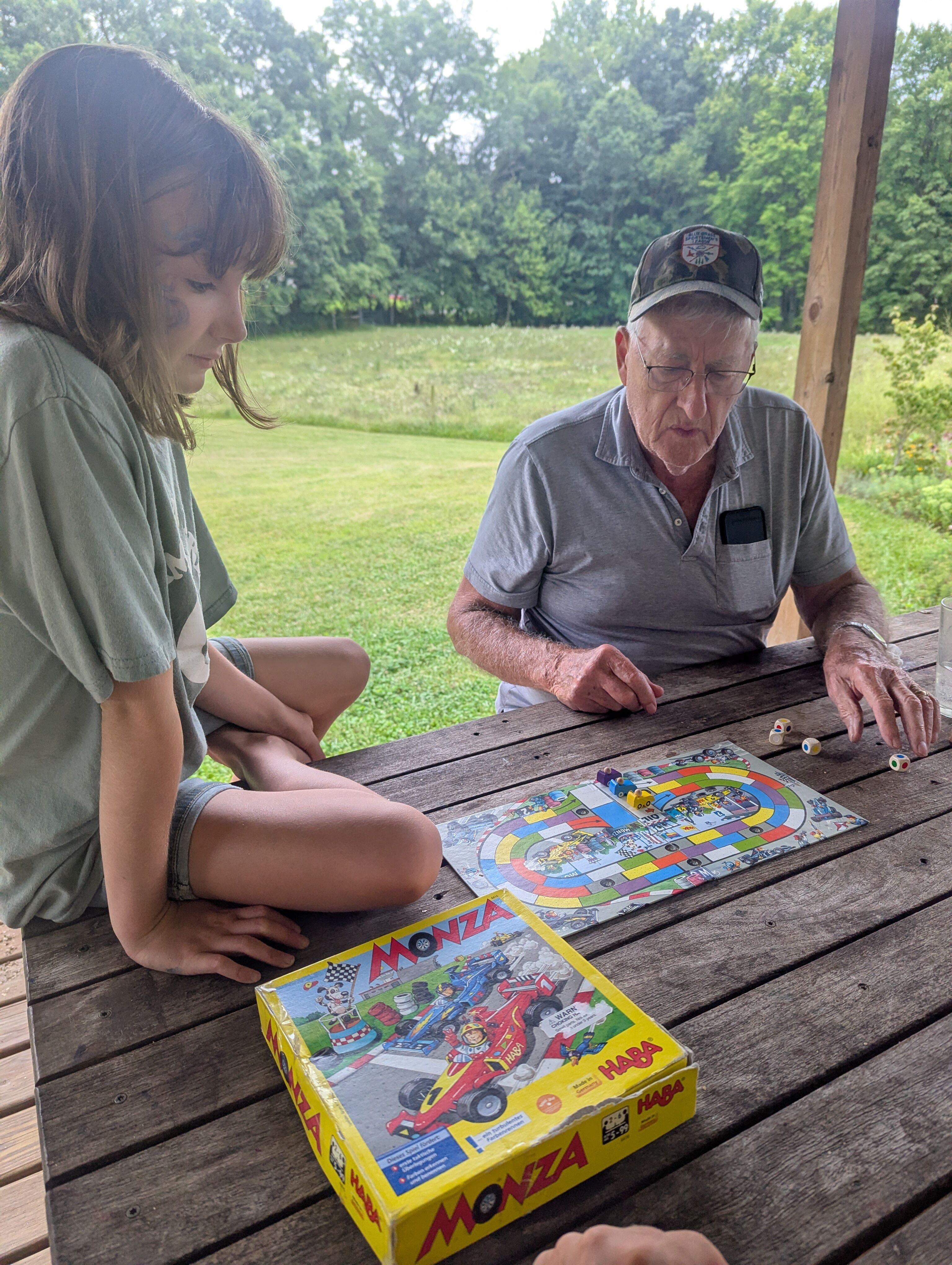 A child and her grandfather playing a small racing board game on a picnic table.