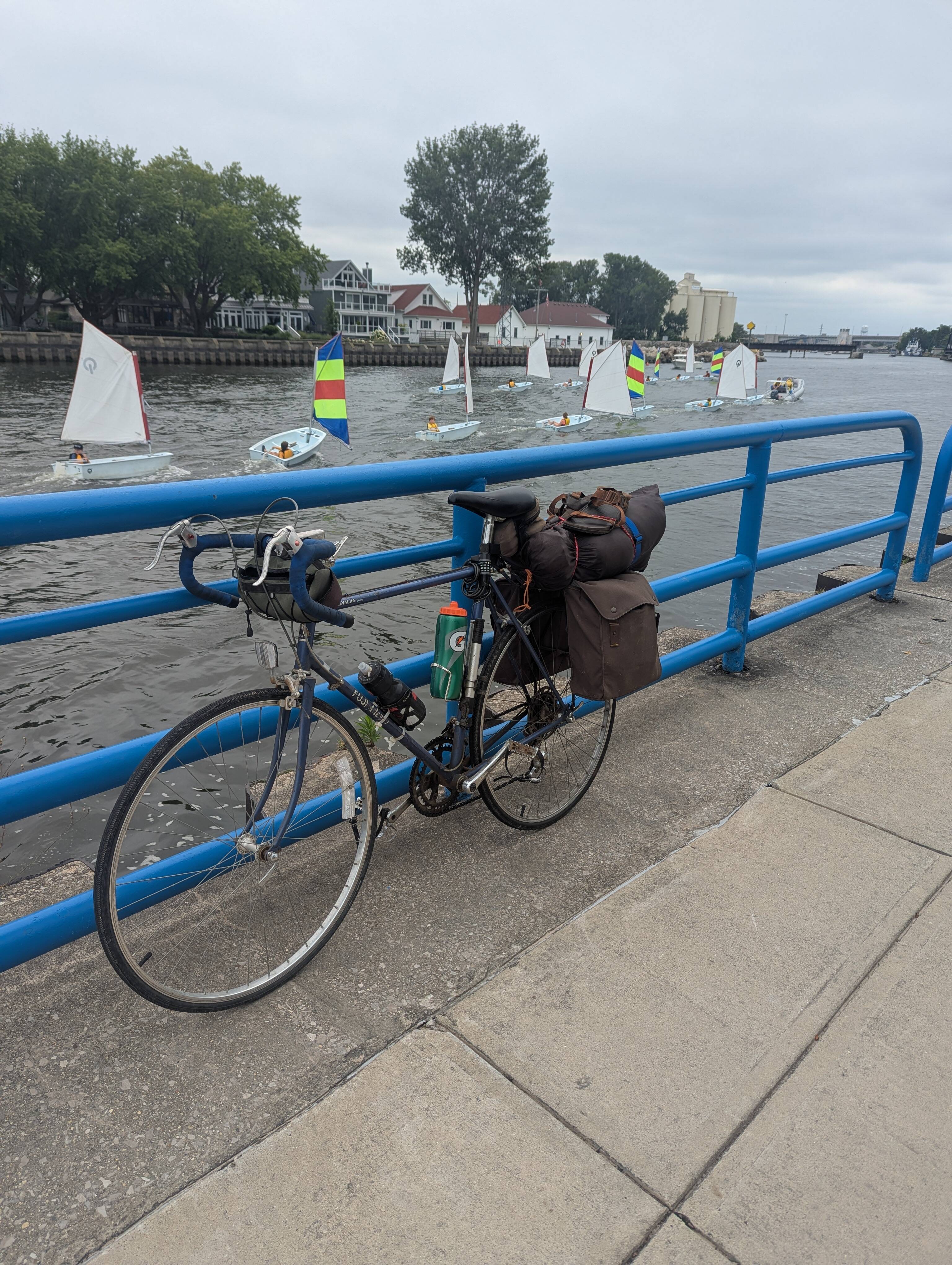 The same loaded bicycle leaning against a railing at a harbor. In the background, several small sailboats with kids are being towed back into the bay by small motorboats.