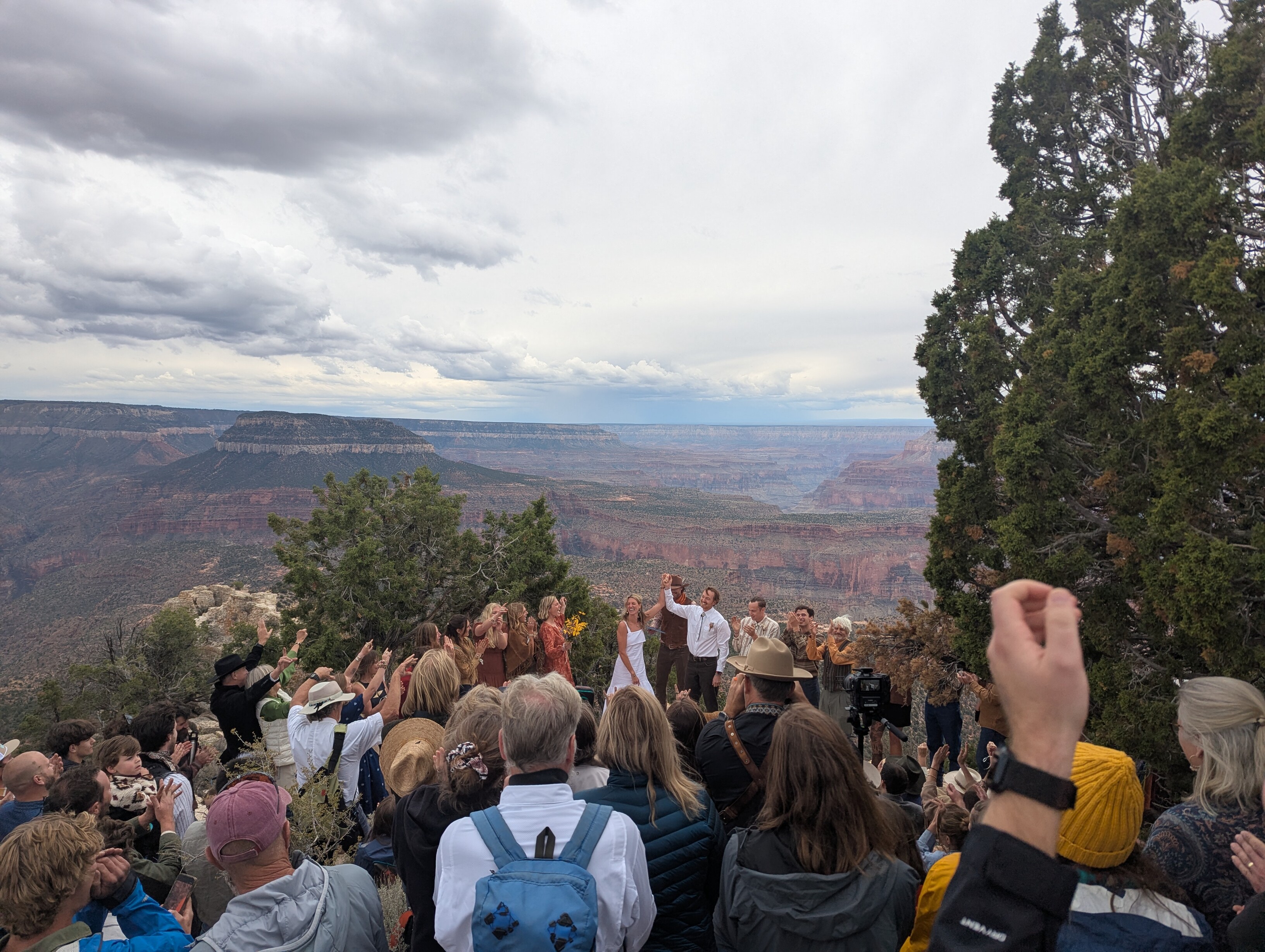 A large group of people on the rim of the Grand Canyon. A bride and groom are in front of the crowd, holding their hands up in the air. The crowd is clapping and cheering for the couple. The canyon stretches off in all directions.