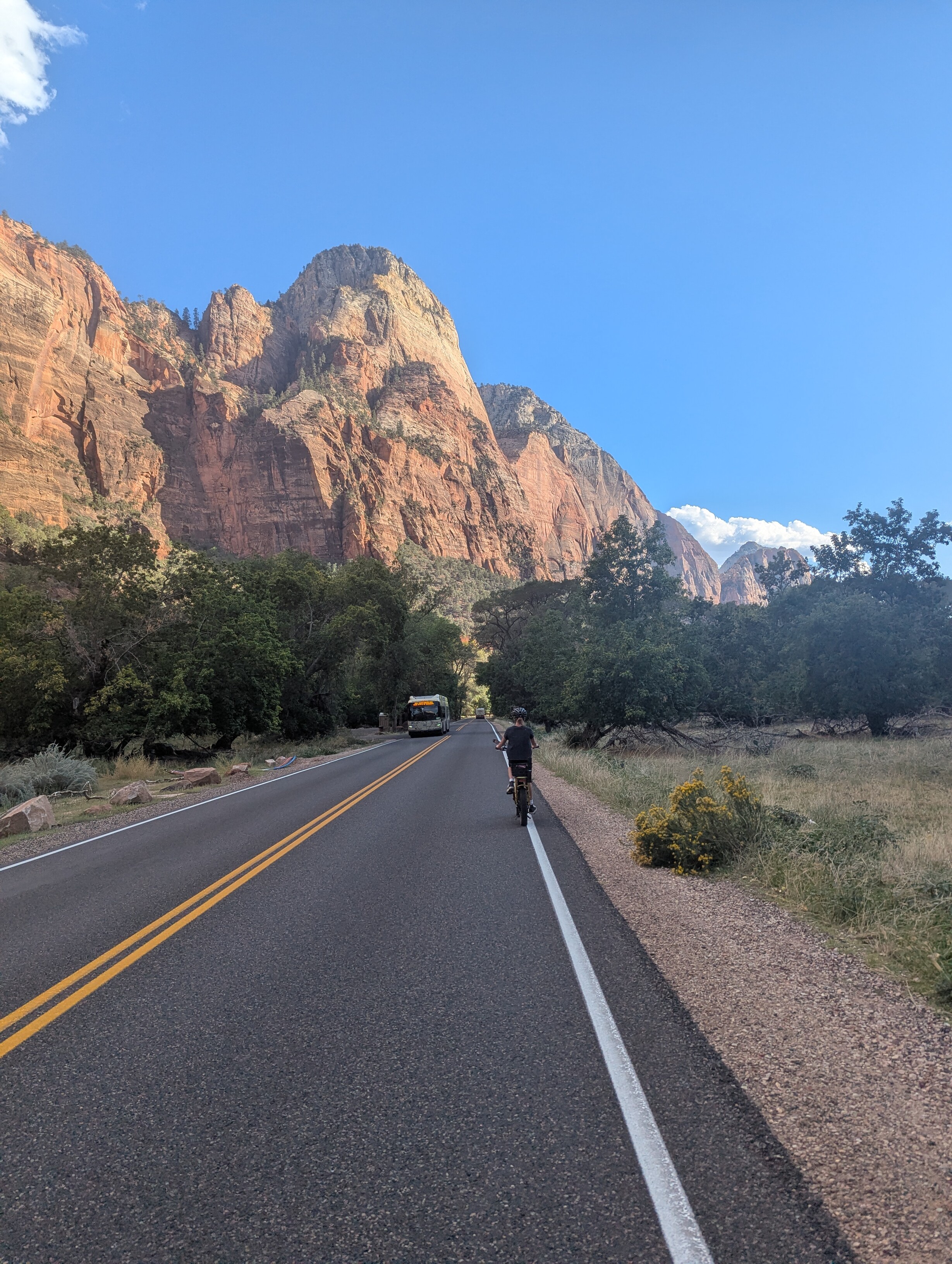 A child on a bike is riding away from the viewer on a road. A shuttle bus is approaching on the opposite side of the road. A large cliff is in bright sunlight in the distance.