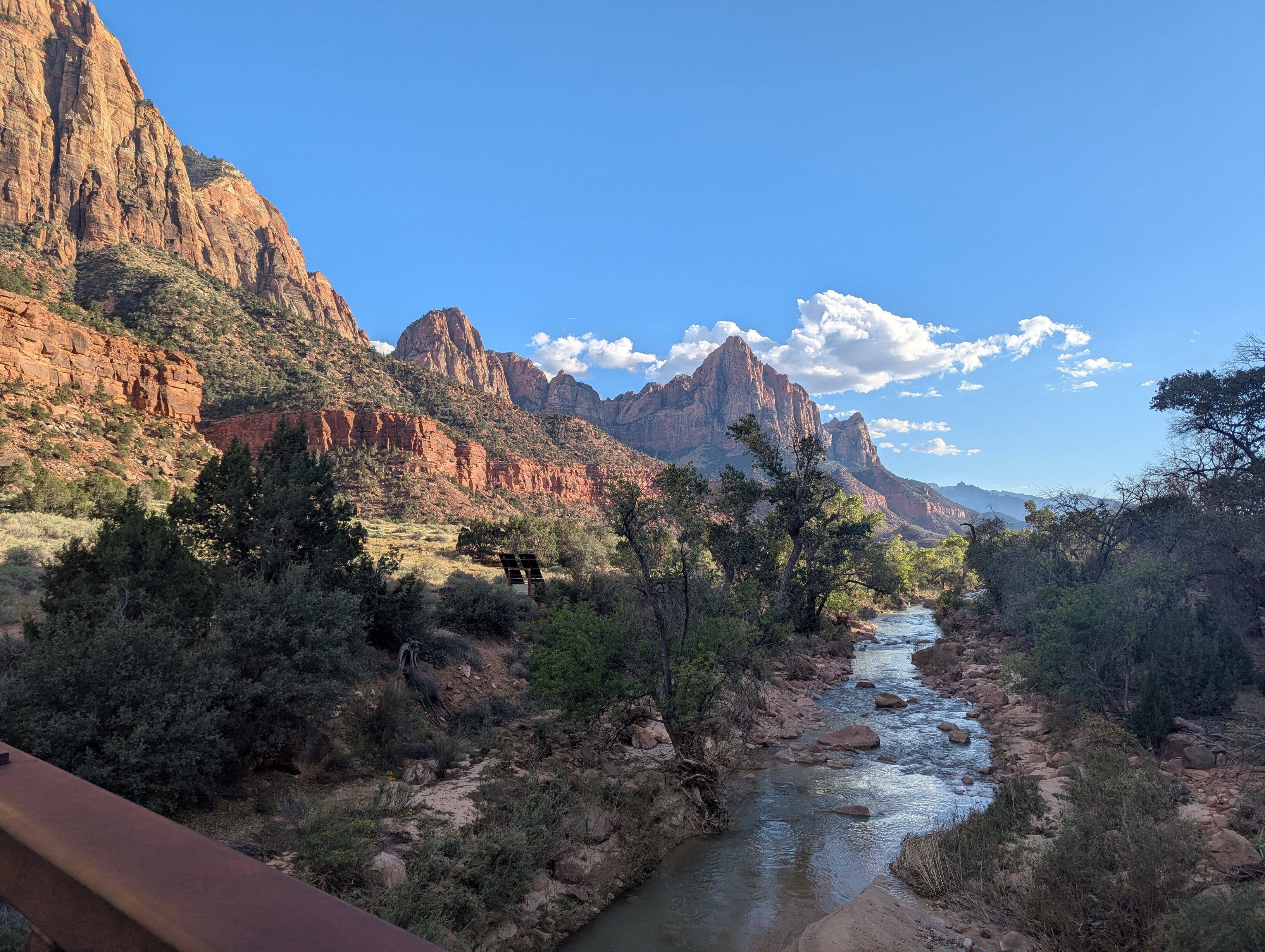 A wide landscape. The Virgin River is below the camera, running peacefully into the distance. The scene is twilight and trees along the river are in shadow. Large rock formations tower in the distance, highlighted yellow and orange in the setting sun.